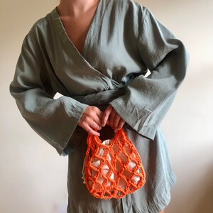 May include: A woman wearing a sage green wrap dress holds a small orange crocheted mesh bag.