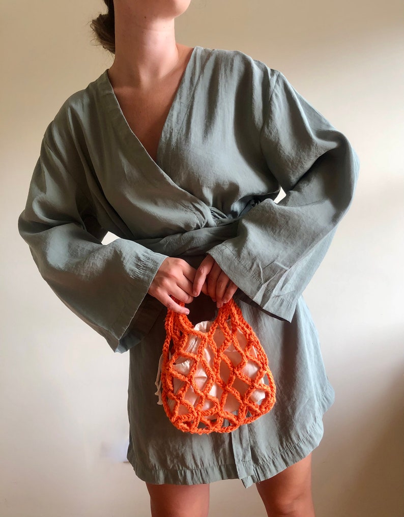 May include: A woman wearing a sage green wrap dress holds a small orange crocheted mesh bag.