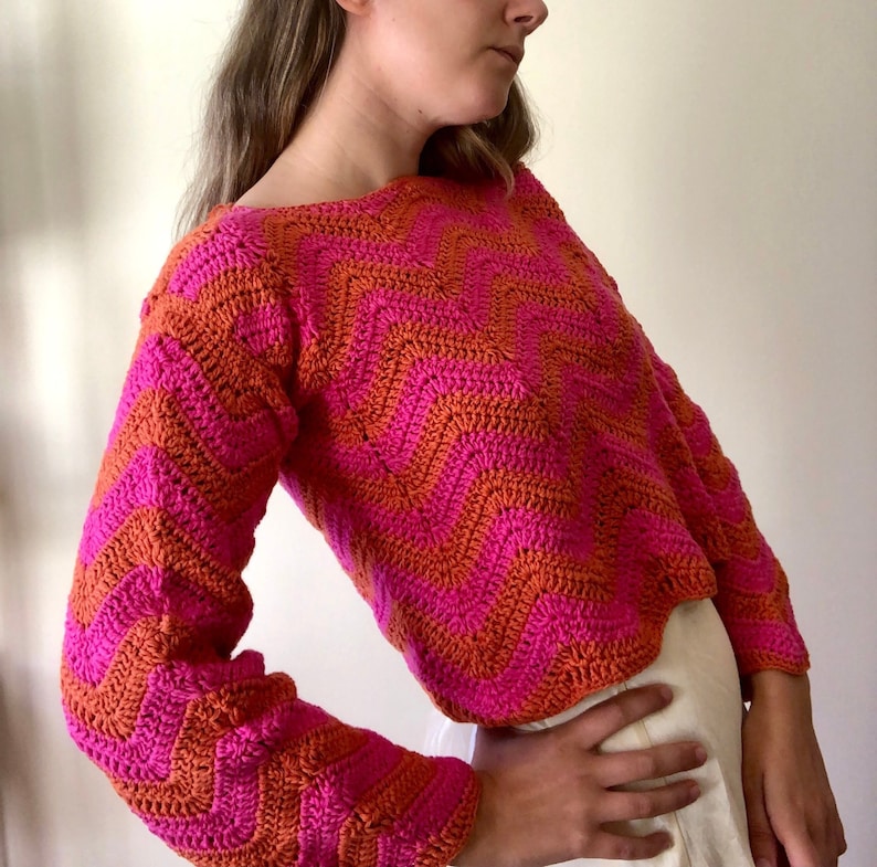 May include: A pink and orange crocheted sweater with a chevron pattern. The sweater has long sleeves and a cropped length.
