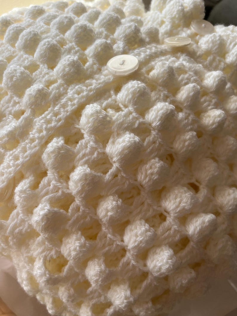 May include: White crocheted fabric with a textured, raised pattern. The fabric has a button in the center.