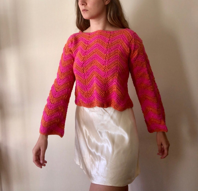 May include: A pink and orange crocheted sweater with a chevron pattern. The sweater has long sleeves and a cropped length.