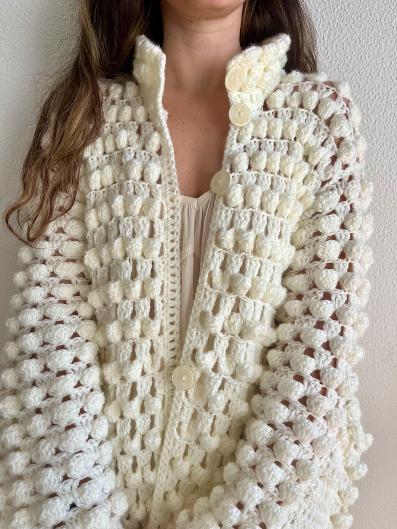 May include: A white crocheted cardigan with a textured, bobble stitch pattern. The cardigan has a high collar and a button closure.