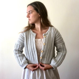 K&ouml;nnte beinhalten: Ein hellgrauer H&auml;kelcardigan mit Knopfleiste und langen &Auml;rmeln. Der Cardigan hat ein zartes Lochmuster.