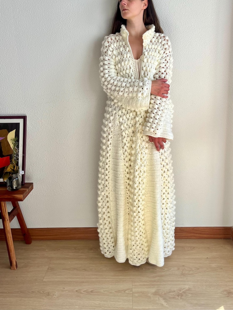May include: A long, white crocheted robe with a textured, bobble stitch pattern. The robe has a high collar, long sleeves, and a full skirt.