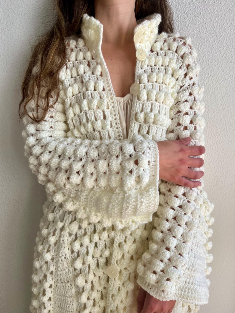 May include: A white crocheted cardigan with a textured bobble stitch pattern. The cardigan has a collar, long sleeves, and a button closure.