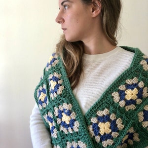 May include: A green, blue, and yellow crocheted shawl with a granny square pattern. The shawl is draped over the shoulders of a person wearing a white long-sleeved shirt.