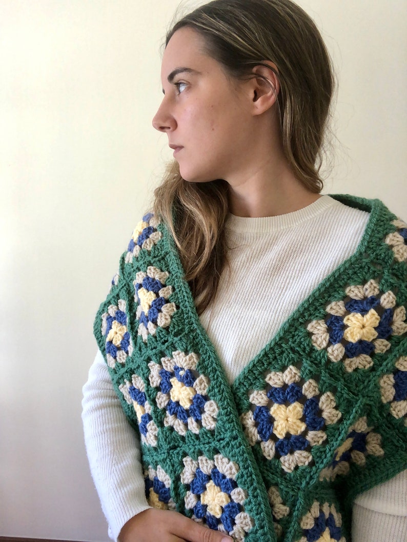 May include: A green, blue, and yellow crocheted shawl with a granny square pattern. The shawl is draped over the shoulders of a person wearing a white long-sleeved shirt.