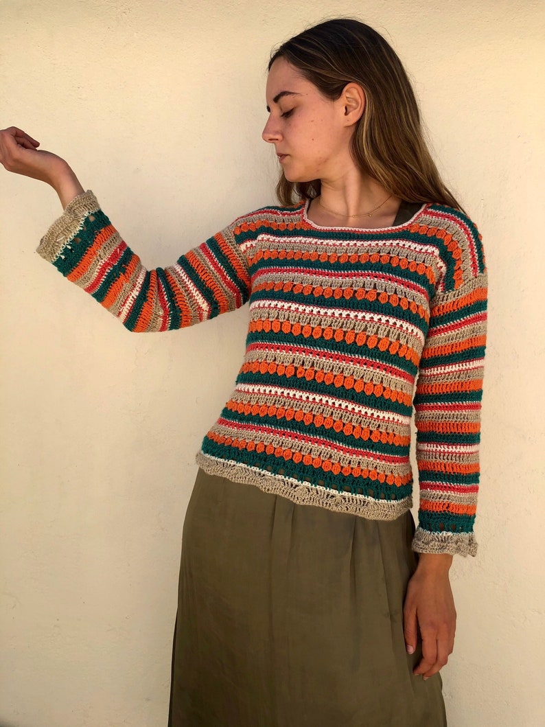 May include: A striped crochet sweater with long sleeves in shades of green, orange, white, and beige. The sweater has a textured design with small, raised circles.