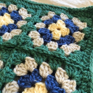 May include: Close-up of a crocheted granny square blanket with a green, beige, blue, and yellow color scheme.