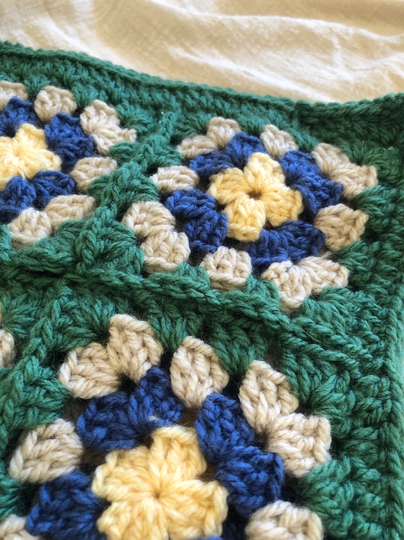 May include: Close-up of a crocheted granny square blanket with a green, beige, blue, and yellow color scheme.