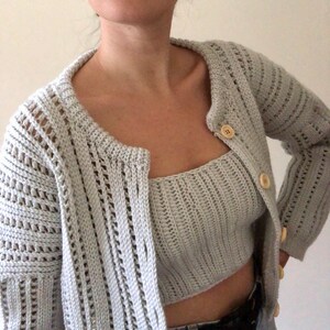 K&ouml;nnte beinhalten: Ein hellgrauer H&auml;kelcardigan mit passendem Crop-Top. Der Cardigan hat einen Knopfverschluss und eine lockere Passform. Das Top ist &auml;rmellos und hat einen geraden Ausschnitt.