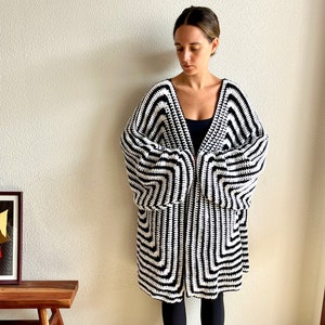May include: A black and white crochet cardigan with a geometric pattern. The long-sleeved cardigan is knee-length and has a V-neck design. The garment is made of a soft, textured yarn and is worn by a person.