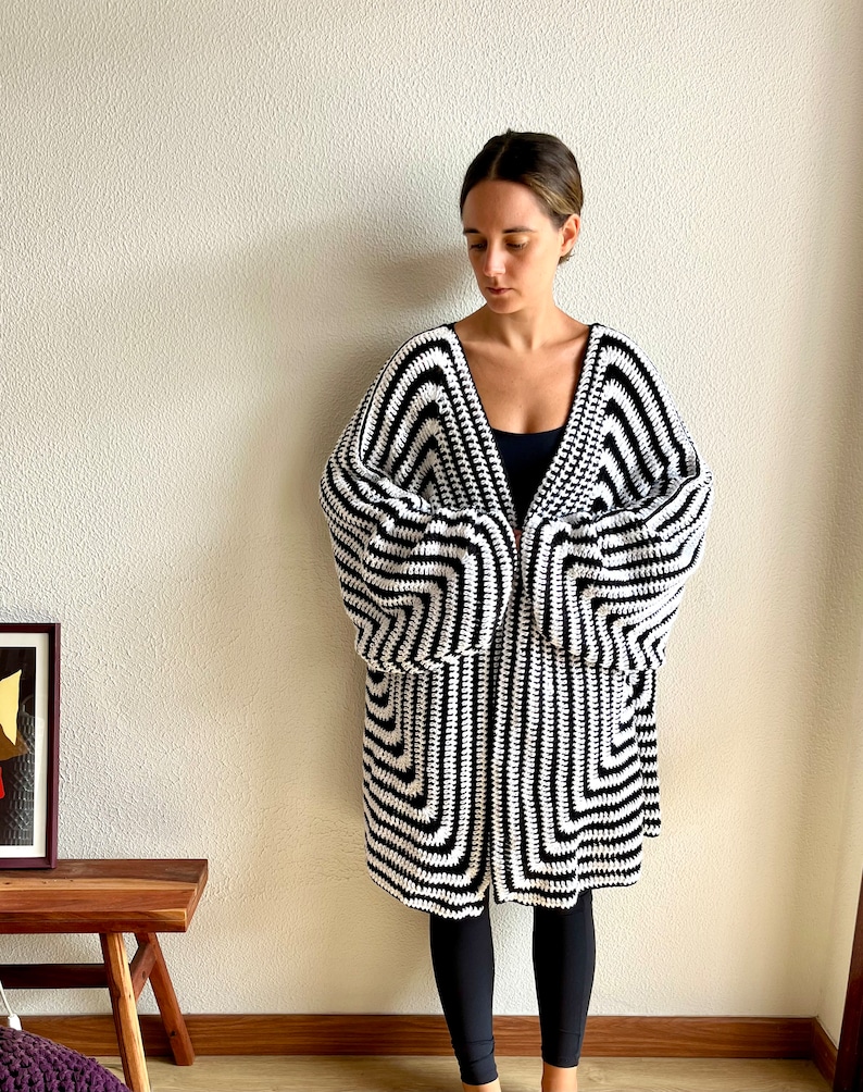 May include: A black and white crochet cardigan with a geometric pattern. The long-sleeved cardigan is knee-length and has a V-neck design. The garment is made of a soft, textured yarn and is worn by a person.