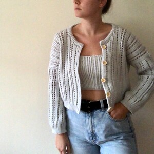 K&ouml;nnte beinhalten: Ein wei&szlig;er H&auml;kelcardigan mit passendem Crop-Top und Kn&ouml;pfen. Der Cardigan hat eine lockere Passform und lange &Auml;rmel. Das Model tr&auml;gt eine blaue Jeans und einen schwarzen G&uuml;rtel.