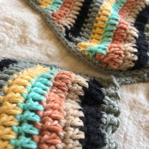Op de afbeelding: Close-up van een gehaakt topje met een gestreept patroon in tinten groen, zwart, beige, geel en lichtblauw.