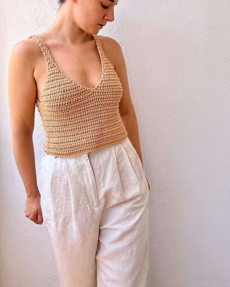 May include: A beige crochet tank top with a v-neckline. The top is worn with white linen pants.