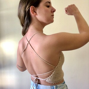 May include: A beige crochet halter top with a criss-cross back and a tassel tie. The top is worn with light blue denim jeans.