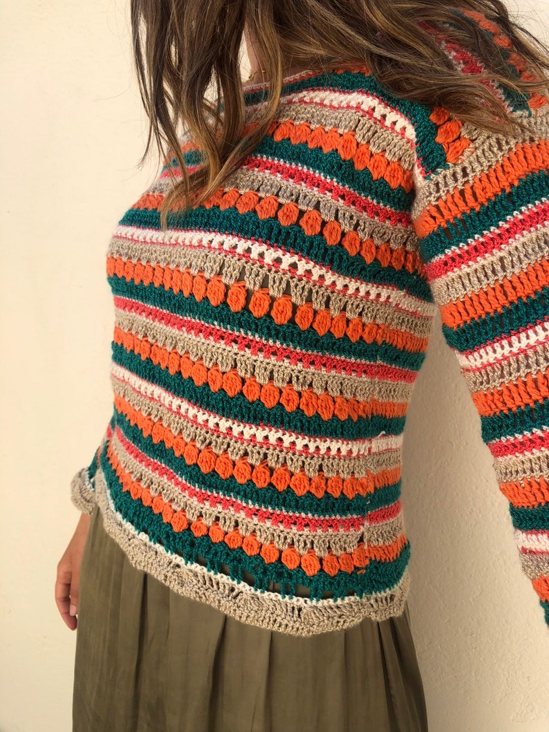 May include: A striped crochet top with long sleeves in shades of orange, green, beige, and pink. The top has a loose fit and a relaxed style.