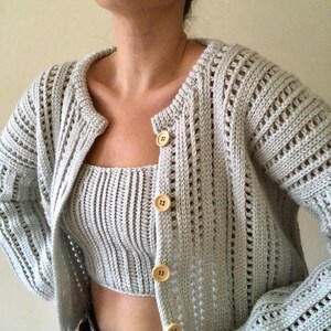 K&ouml;nnte beinhalten: Ein hellgrauer Strickcardigan mit passendem Crop-Top. Der Cardigan hat einen Knopfverschluss und ein zartes Lochmuster. Das Top hat einen runden Ausschnitt und eine gerippte Struktur.