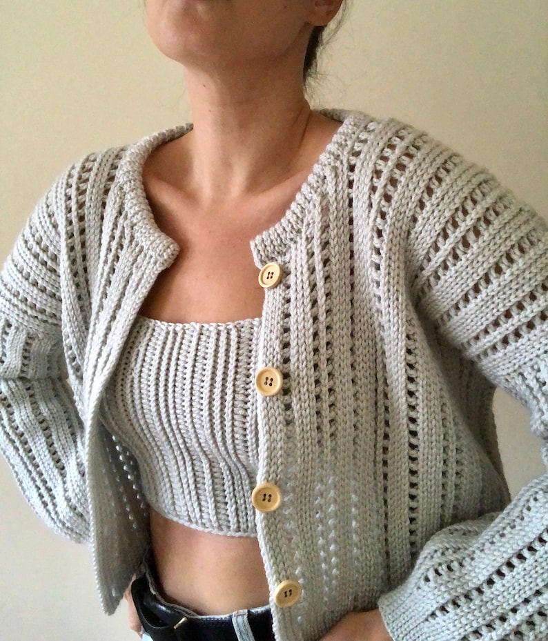 K&ouml;nnte beinhalten: Ein hellgrauer Strickcardigan mit passendem Crop-Top. Der Cardigan hat einen Knopfverschluss und ein zartes Lochmuster. Das Top hat einen runden Ausschnitt und eine gerippte Struktur.