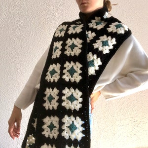 May include: A black and white crocheted scarf with green accents. The scarf is made of granny squares and is worn around the neck.