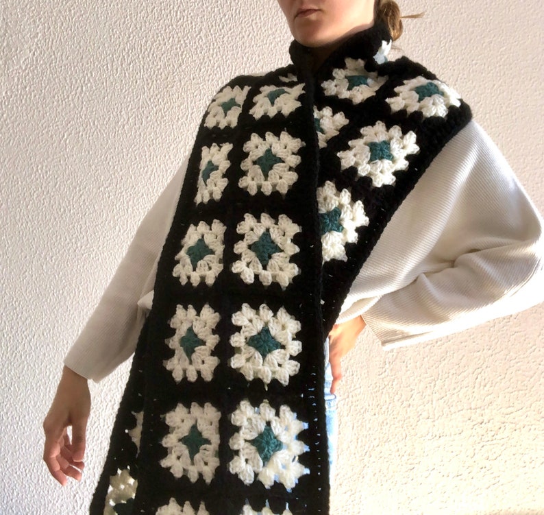 May include: A black and white crocheted scarf with green accents. The scarf is made of granny squares and is worn around the neck.