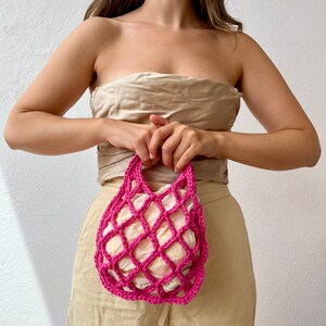 May include: A bright pink crocheted mesh bag with a white fabric lining. The bag is held by a person wearing a beige top and pants.