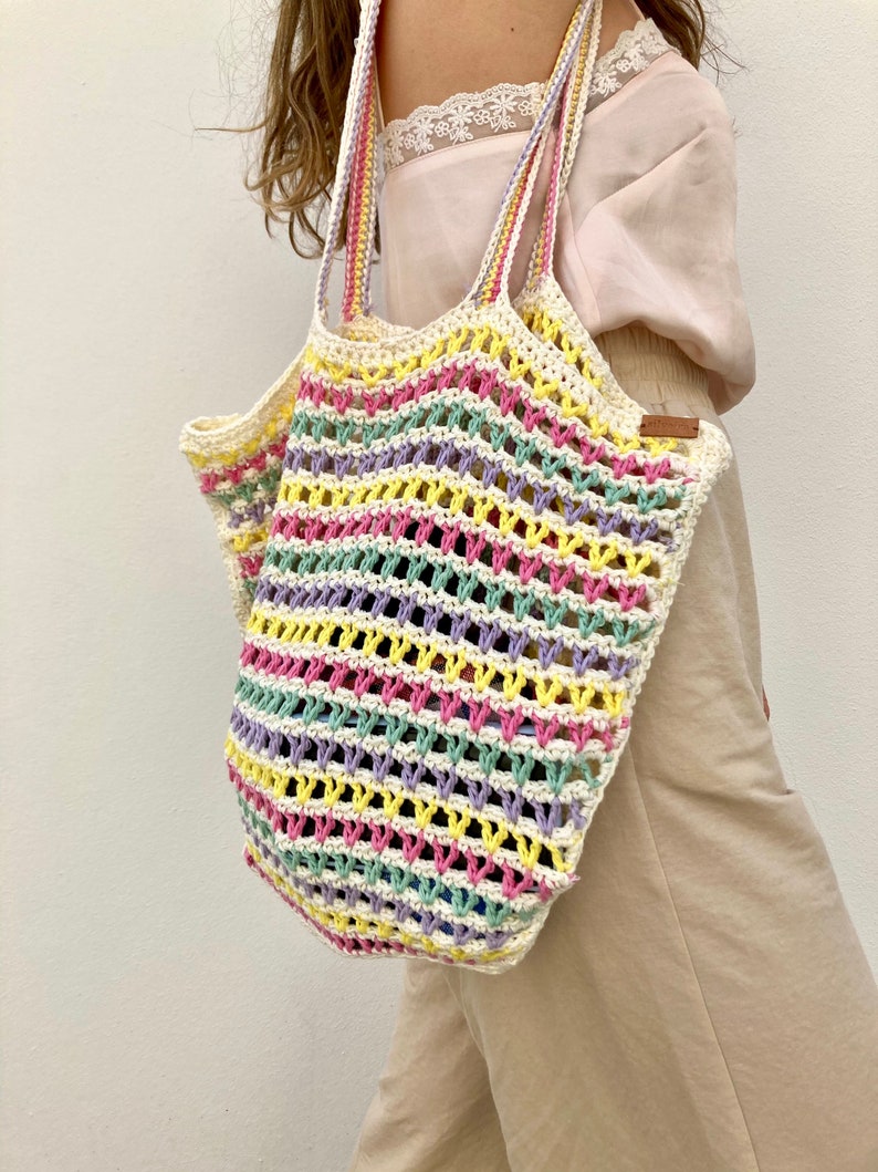 May include: A colorful crocheted tote bag with a striped pattern in pink, yellow, green, and purple. The bag is made of a lightweight, breathable material and has a wide, comfortable handle.