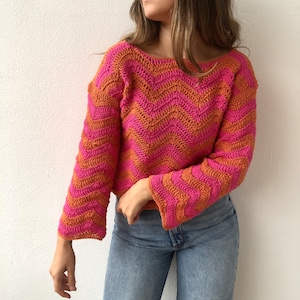 May include: A pink and orange crocheted sweater with a chevron pattern. The sweater has long sleeves and a relaxed fit.