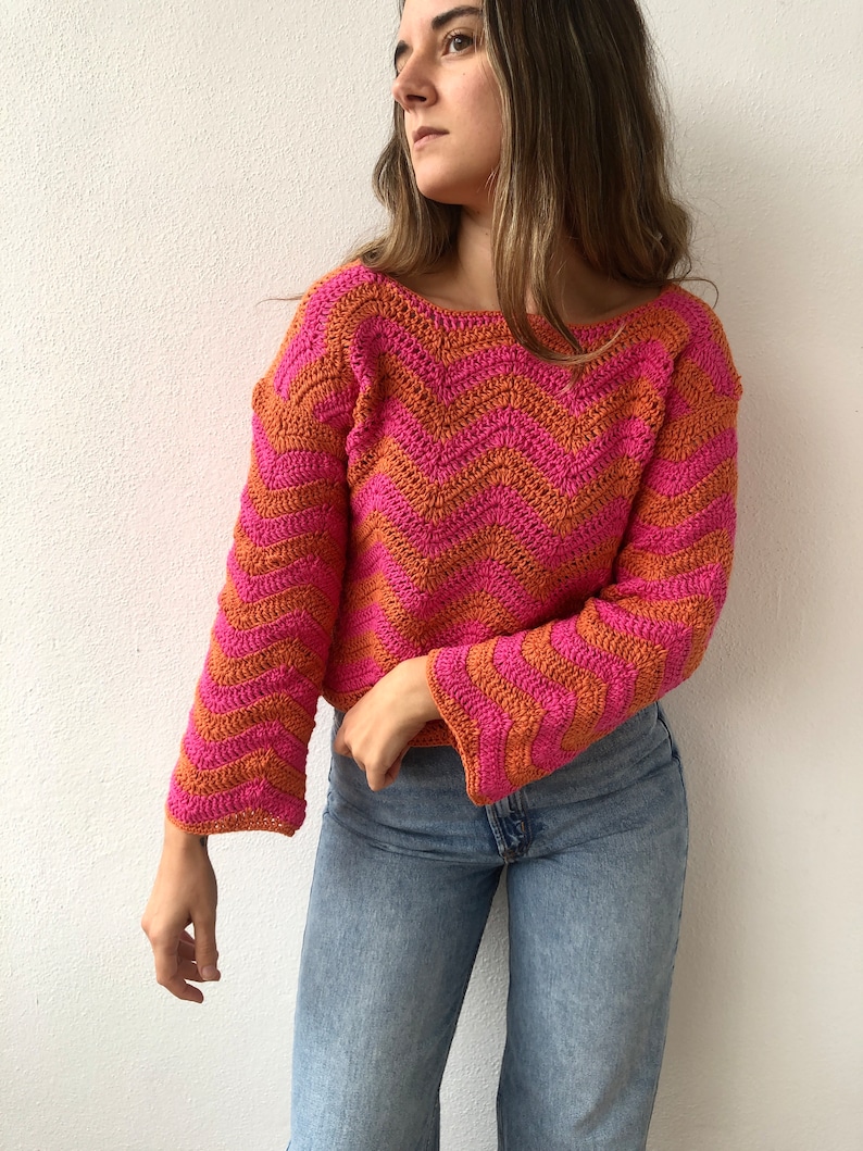 May include: A pink and orange crocheted sweater with a chevron pattern. The sweater has long sleeves and a relaxed fit.