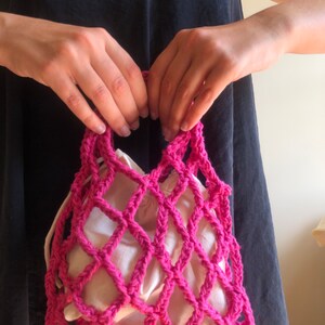 May include: A pink crocheted mesh bag with a white fabric lining. The bag is held by a person's hands.
