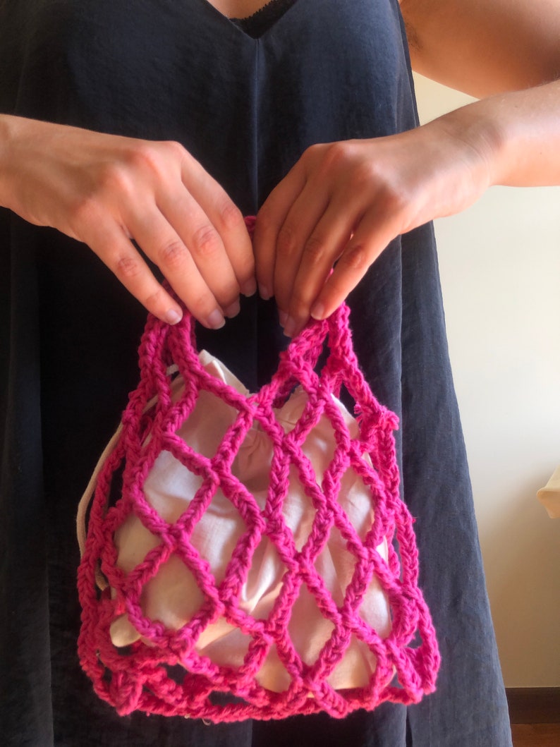 May include: A pink crocheted mesh bag with a white fabric lining. The bag is held by a person's hands.