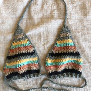 Op de afbeelding: Een gehaakt bikini-topje met een gestreept patroon in tinten zwart, grijs, geel, roze en blauw. Het topje heeft verstelbare bandjes en strikjes aan de achterkant.