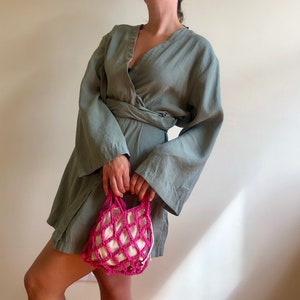May include: A woman wearing a sage green wrap dress with long sleeves and a pink crocheted mesh bag.