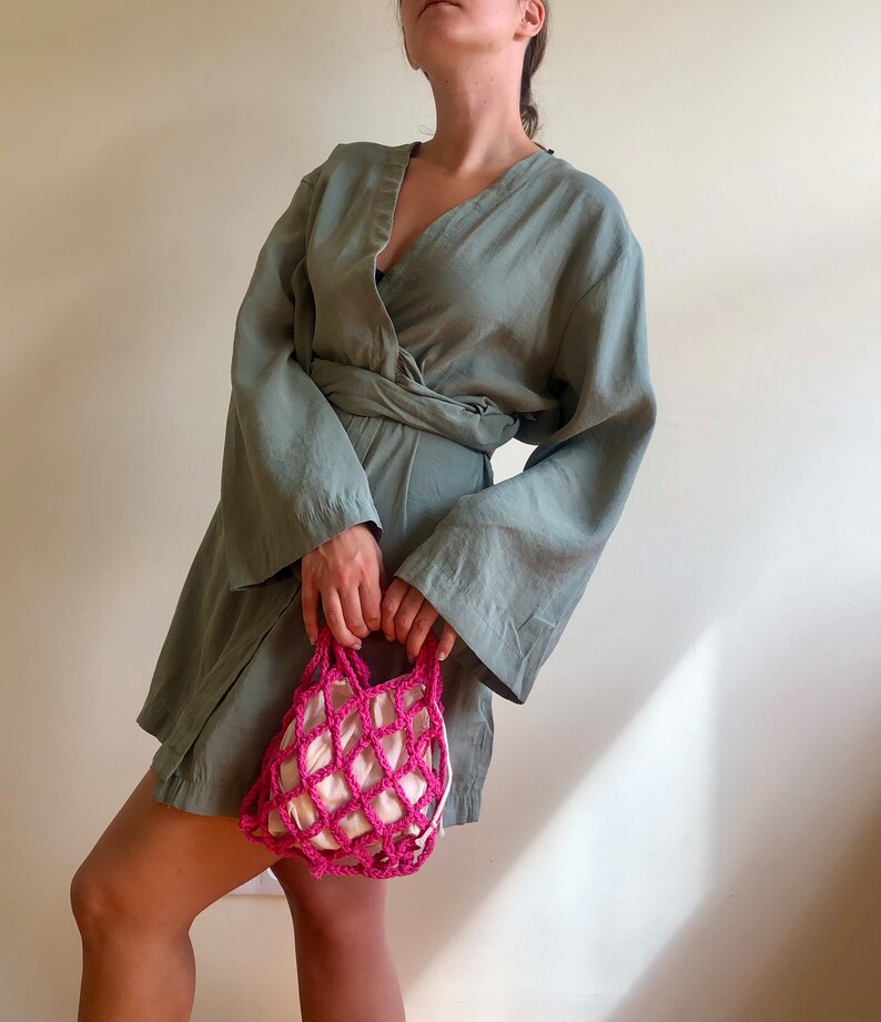 May include: A woman wearing a sage green wrap dress with long sleeves and a pink crocheted mesh bag.