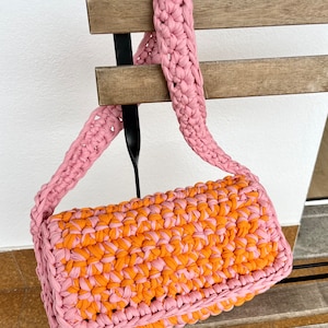 May include: A pink and orange crocheted handbag with a long pink strap. The bag is hanging from a wooden chair.
