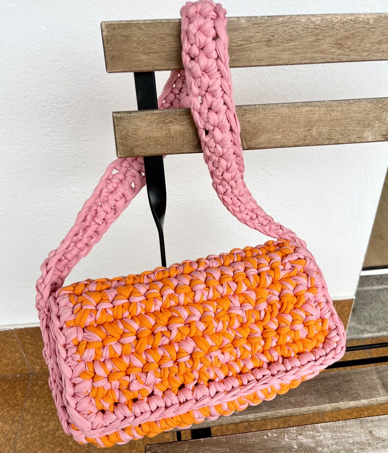 May include: A pink and orange crocheted handbag with a long pink strap. The bag is hanging from a wooden chair.
