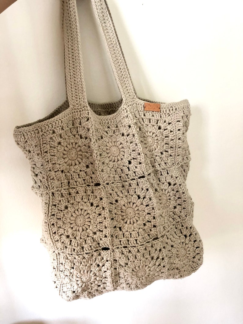 May include: A beige crocheted tote bag with a brown leather tag. The bag is made of a square pattern with a circular design in the center of each square.