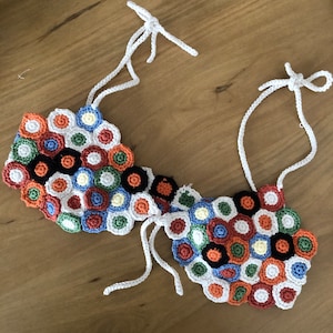 Puede incluir: Un top de crochet con un patrón geométrico colorido en tonos de naranja, verde, azul, blanco y negro. El top tiene lazos blancos que se pueden ajustar para un ajuste personalizado.