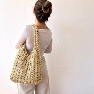 K&ouml;nnte beinhalten: Eine beige geh&auml;kelte Tragetasche mit einer breiten, offenen Oberseite und einem einzigen Riemen. Die Tasche wird &uuml;ber der Schulter einer Person getragen, die ein wei&szlig;es Langarmshirt und eine beige Hose tr&auml;gt.