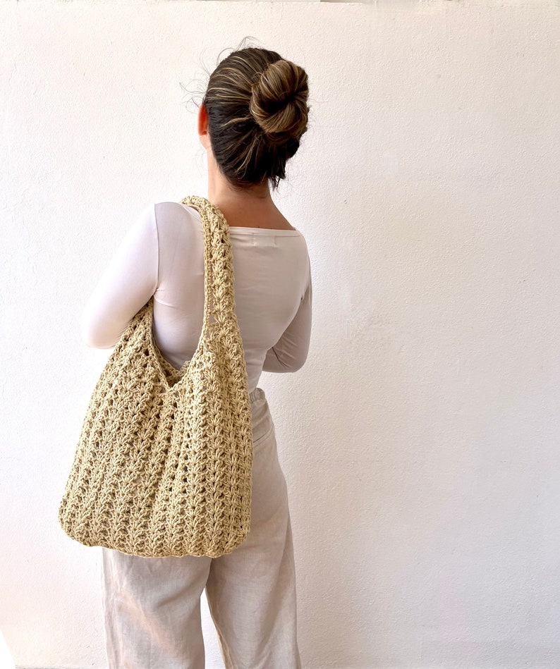 K&ouml;nnte beinhalten: Eine beige geh&auml;kelte Tragetasche mit einer breiten, offenen Oberseite und einem einzigen Riemen. Die Tasche wird &uuml;ber der Schulter einer Person getragen, die ein wei&szlig;es Langarmshirt und eine beige Hose tr&auml;gt.