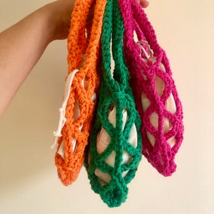 May include: Three crocheted string bags in orange, green, and pink. The bags are hanging from a hand and are made with a simple, open weave pattern.