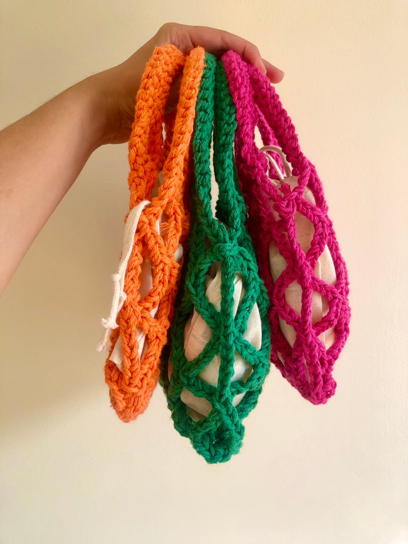 May include: Three crocheted string bags in orange, green, and pink. The bags are hanging from a hand and are made with a simple, open weave pattern.