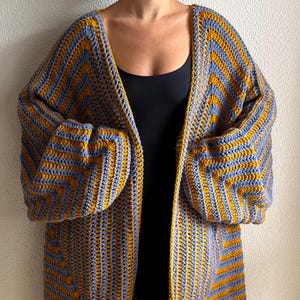 May include: A handmade, open-front crochet cardigan in a chevron pattern. The long, oversized sweater features alternating stripes of mustard yellow and gray-blue yarn. The sleeves are long and voluminous, and the cardigan falls to mid-thigh.