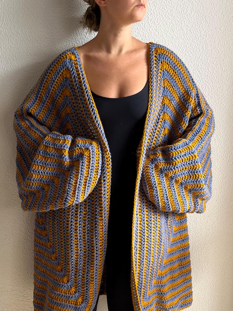 May include: A handmade, open-front crochet cardigan in a chevron pattern. The long, oversized sweater features alternating stripes of mustard yellow and gray-blue yarn. The sleeves are long and voluminous, and the cardigan falls to mid-thigh.