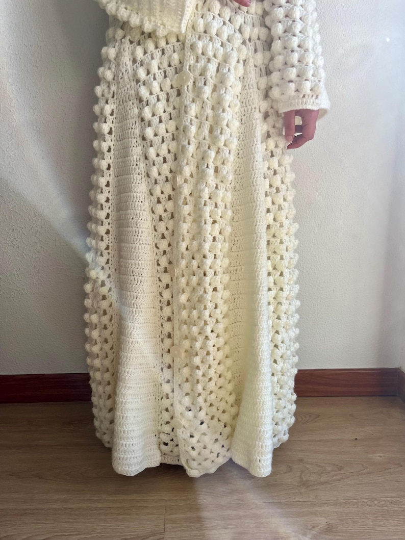 May include: A white crocheted maxi skirt with a textured bobble stitch pattern. The skirt has a flared silhouette and a fitted waistband.