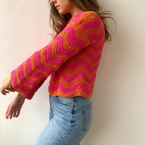May include: A pink and orange crocheted sweater with a chevron pattern. The sweater has long sleeves and a cropped length.
