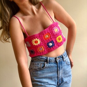 May include: A pink crochet top with colorful flower designs. The top has thin straps and a square neckline.