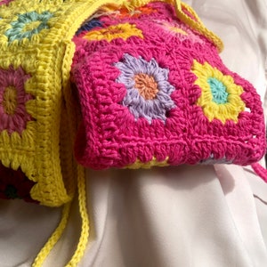 K&ouml;nnte beinhalten: Eine handgefertigte H&auml;keltasche mit floralem Design. Die Tasche ist haupts&auml;chlich pink mit gelben und mehrfarbigen Blumenapplikationen. Der Tr&auml;ger ist gelb. Die Tasche ist aus Garn gefertigt.