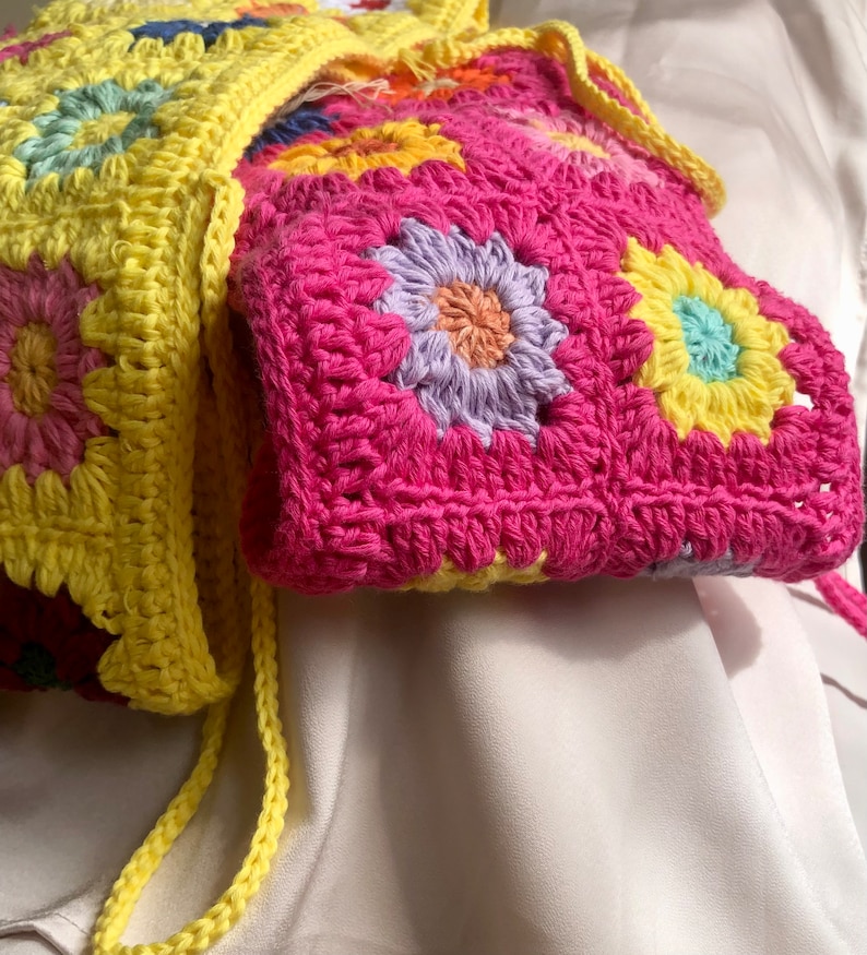 K&ouml;nnte beinhalten: Eine handgefertigte H&auml;keltasche mit floralem Design. Die Tasche ist haupts&auml;chlich pink mit gelben und mehrfarbigen Blumenapplikationen. Der Tr&auml;ger ist gelb. Die Tasche ist aus Garn gefertigt.
